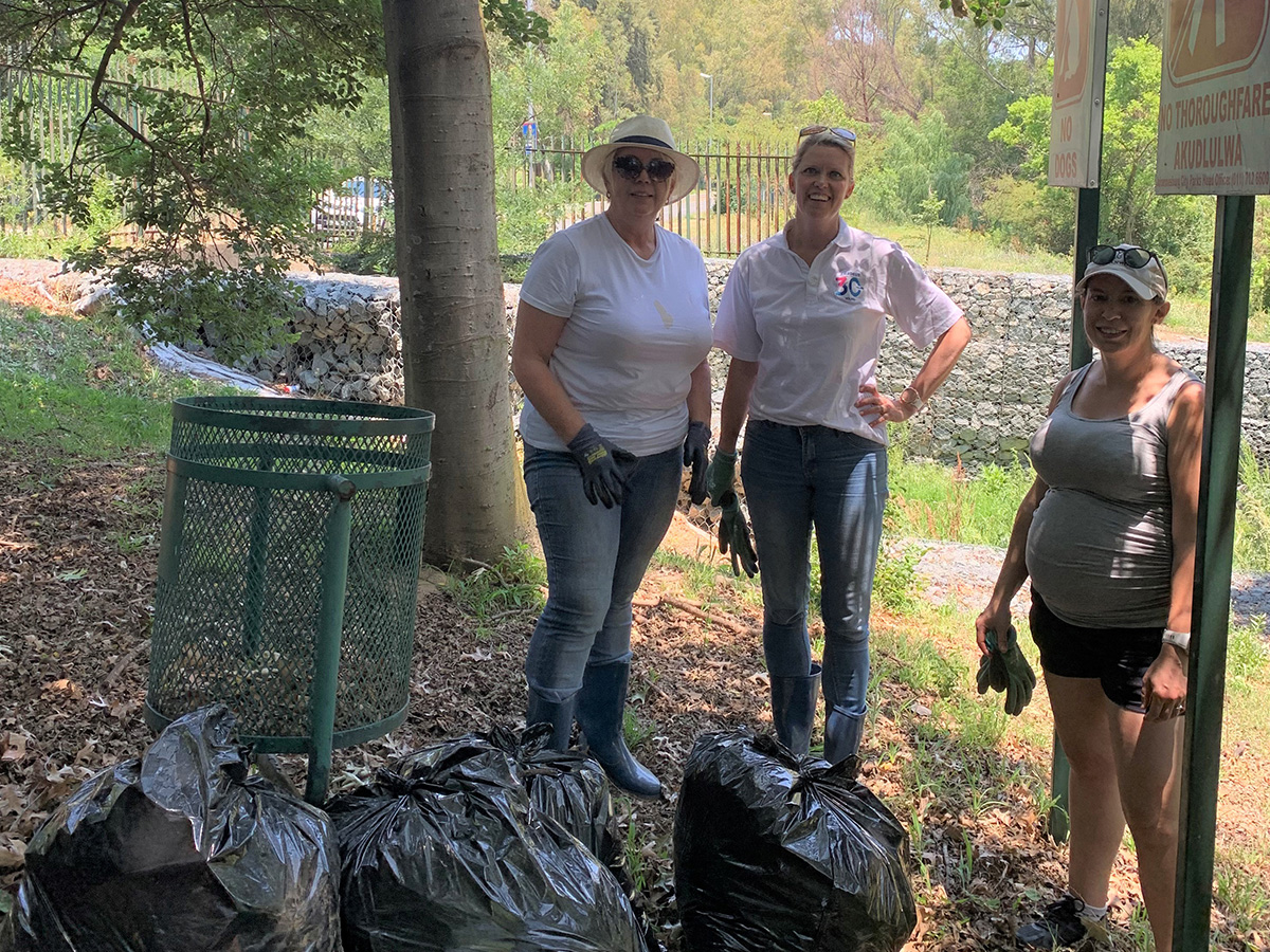 Three women with work clothes and gloves pose in the field together with multiple bags of rubbish. 